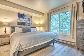 A bedroom with a large bed, a nightstand with a lamp, a dresser, and a window with curtains.