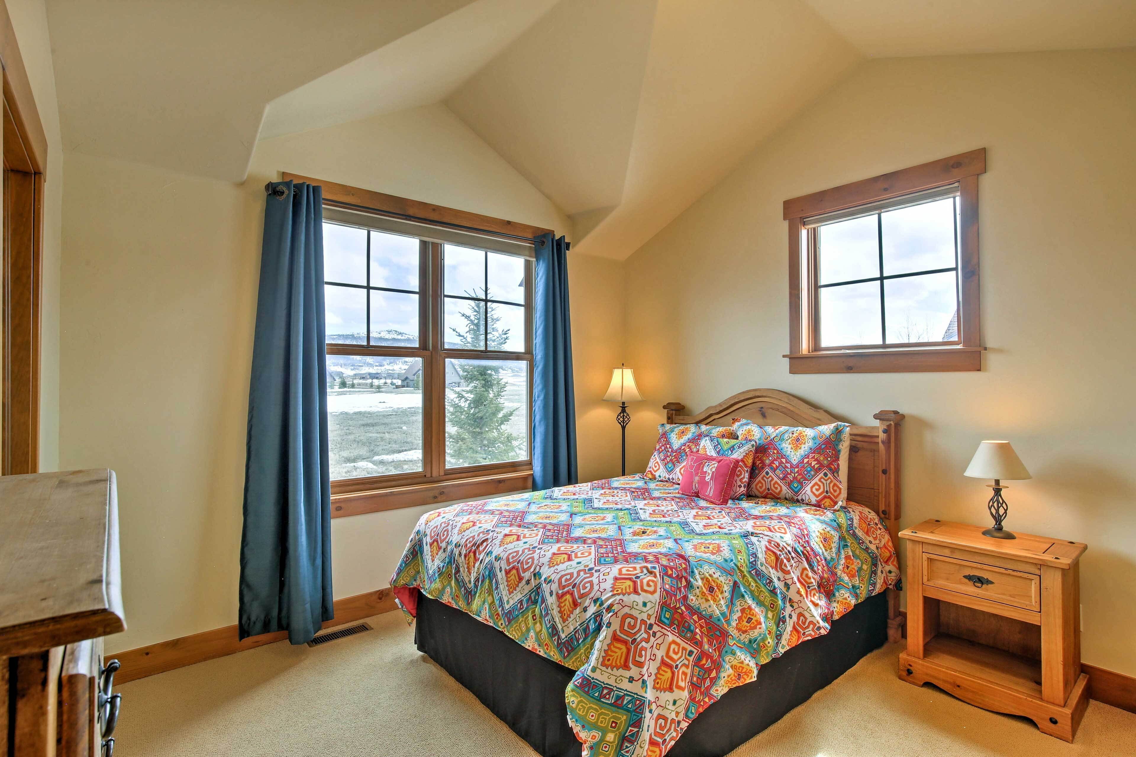 A bedroom with a large bed, a nightstand with a lamp, a window with curtains, and a view of the outdoors.