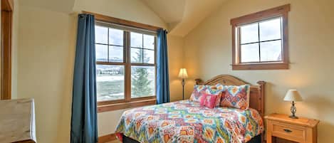 A bedroom with a large bed, a nightstand with a lamp, a window with curtains, and a view of the outdoors.