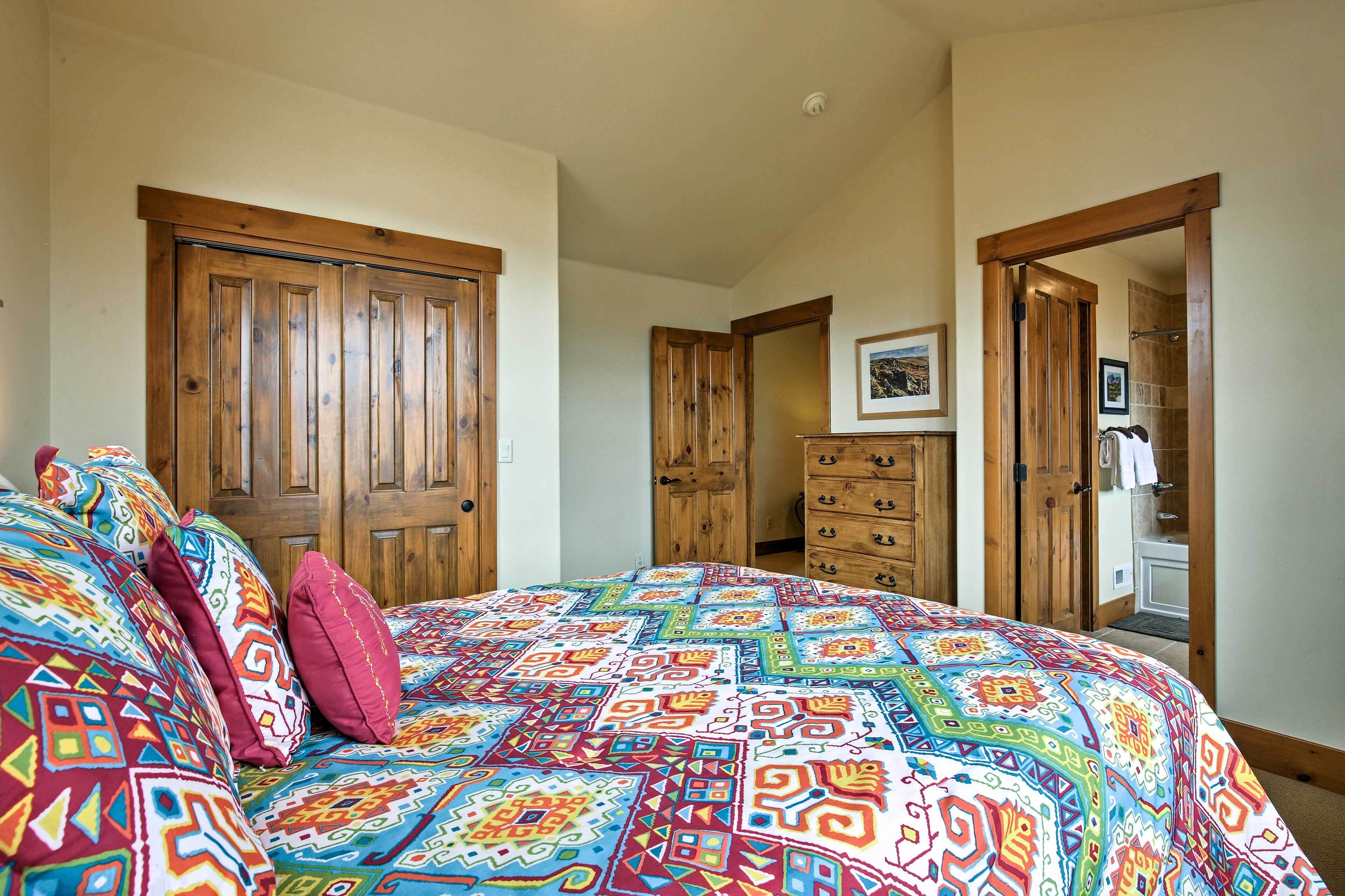 Quarto com colcha colorida, guarda-roupa de madeira e banheiro visível através de uma porta aberta.