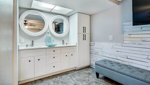 Een badkamer met een wit wastafelmeubel, twee ronde spiegels, een grijze bank en een wandgemonteerde televisie.