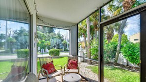 Une terrasse ensoleillée avec une table et deux chaises, donnant sur un jardin bien entretenu, agrémenté de palmiers et d’une pelouse.