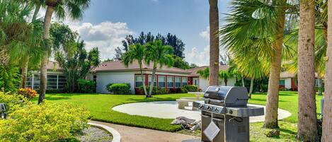 A backyard with a barbecue grill, palm trees, and a house with a red roof.