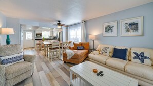 Un salon avec un canapé, un fauteuil et une table basse, un coin repas avec une table et des chaises, et une cuisine à l’arrière-plan.
