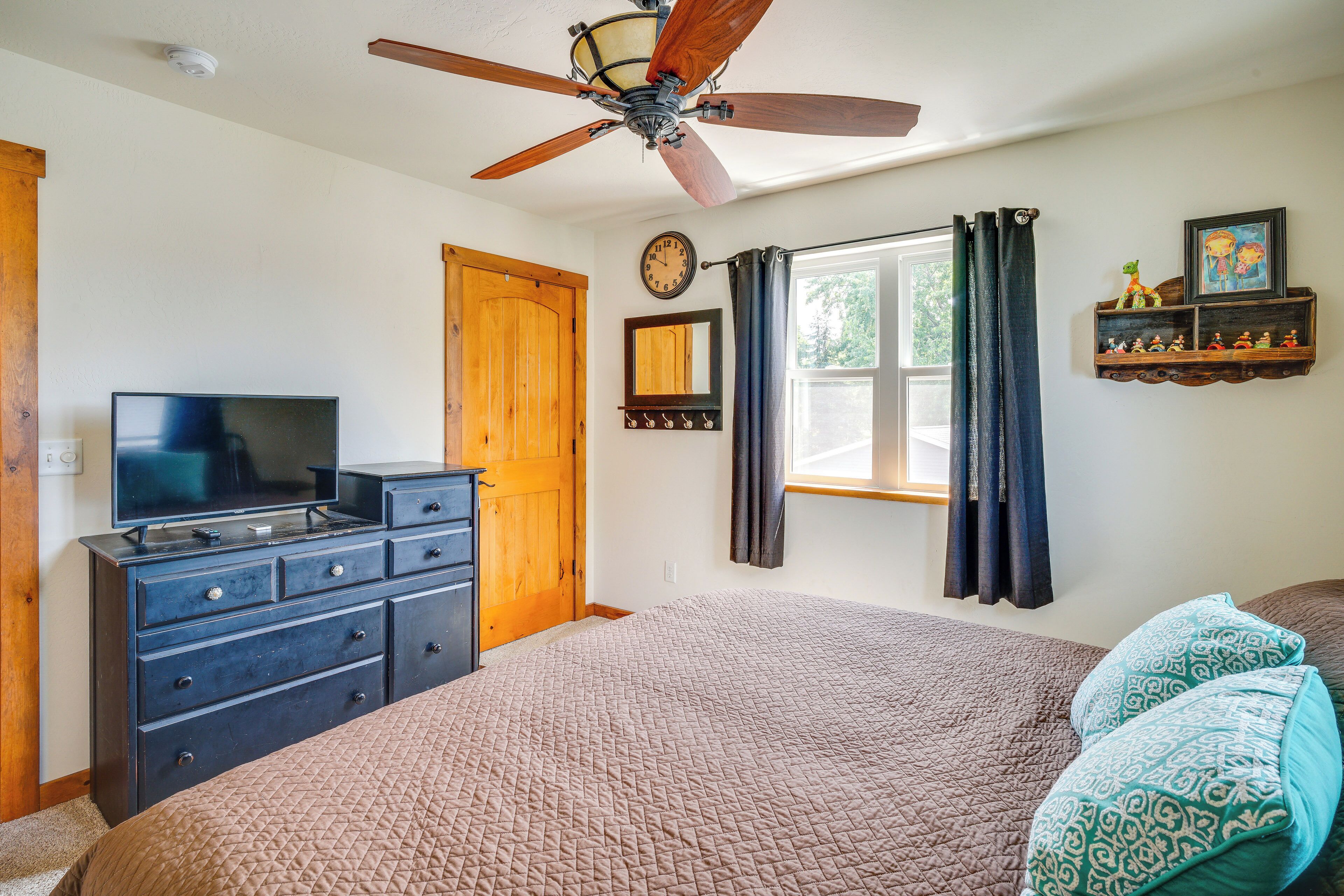 Une chambre à coucher comprenant un lit, une commode, un téléviseur, un ventilateur de plafond, une fenêtre avec des rideaux et des étagères fixées au mur.