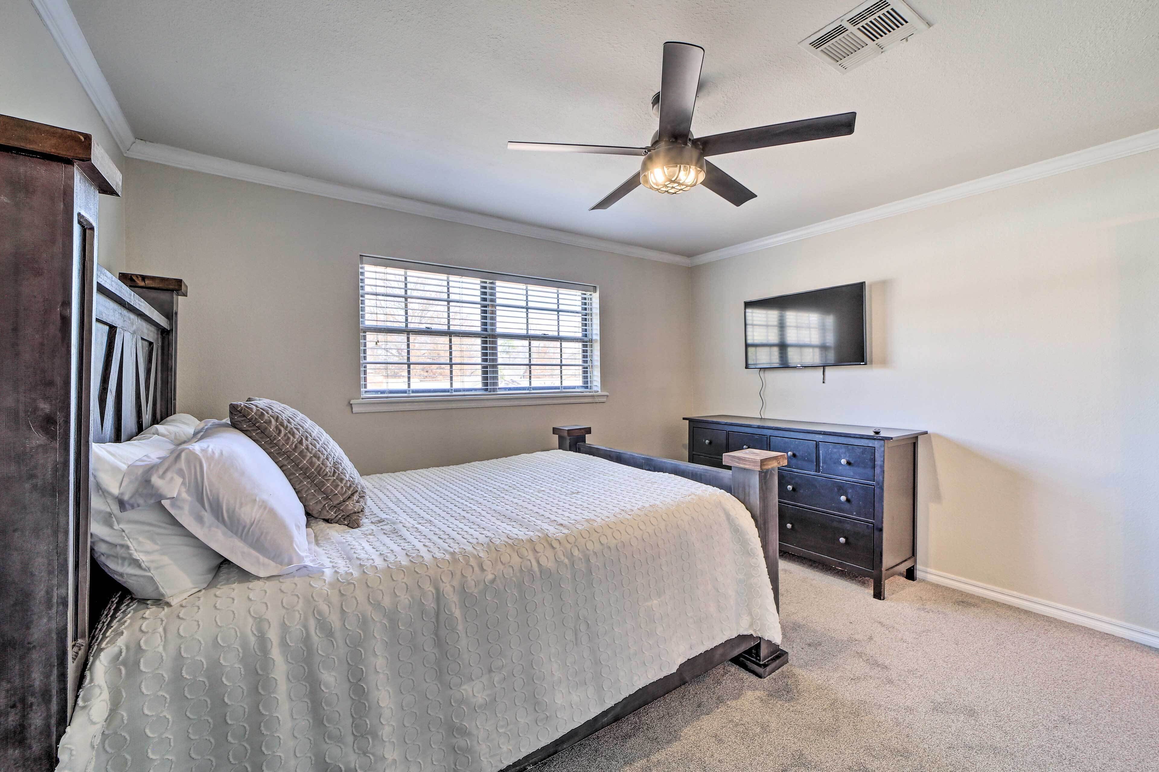 A bedroom with a bed, a ceiling fan, a window, a dresser, and a TV mounted on the wall.
