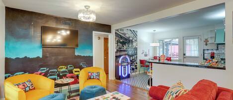 A modern living room with vibrant furniture, a retro jukebox, and a kitchen visible through an open doorway.