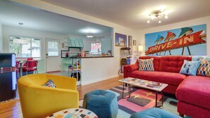 Ein modernes Wohnzimmer mit einem roten Sofa, einem gelben Sessel und einem blauen Hocker. Im Hintergrund ist eine Küche erkennbar, mit einem 'Drive-In' Schild.