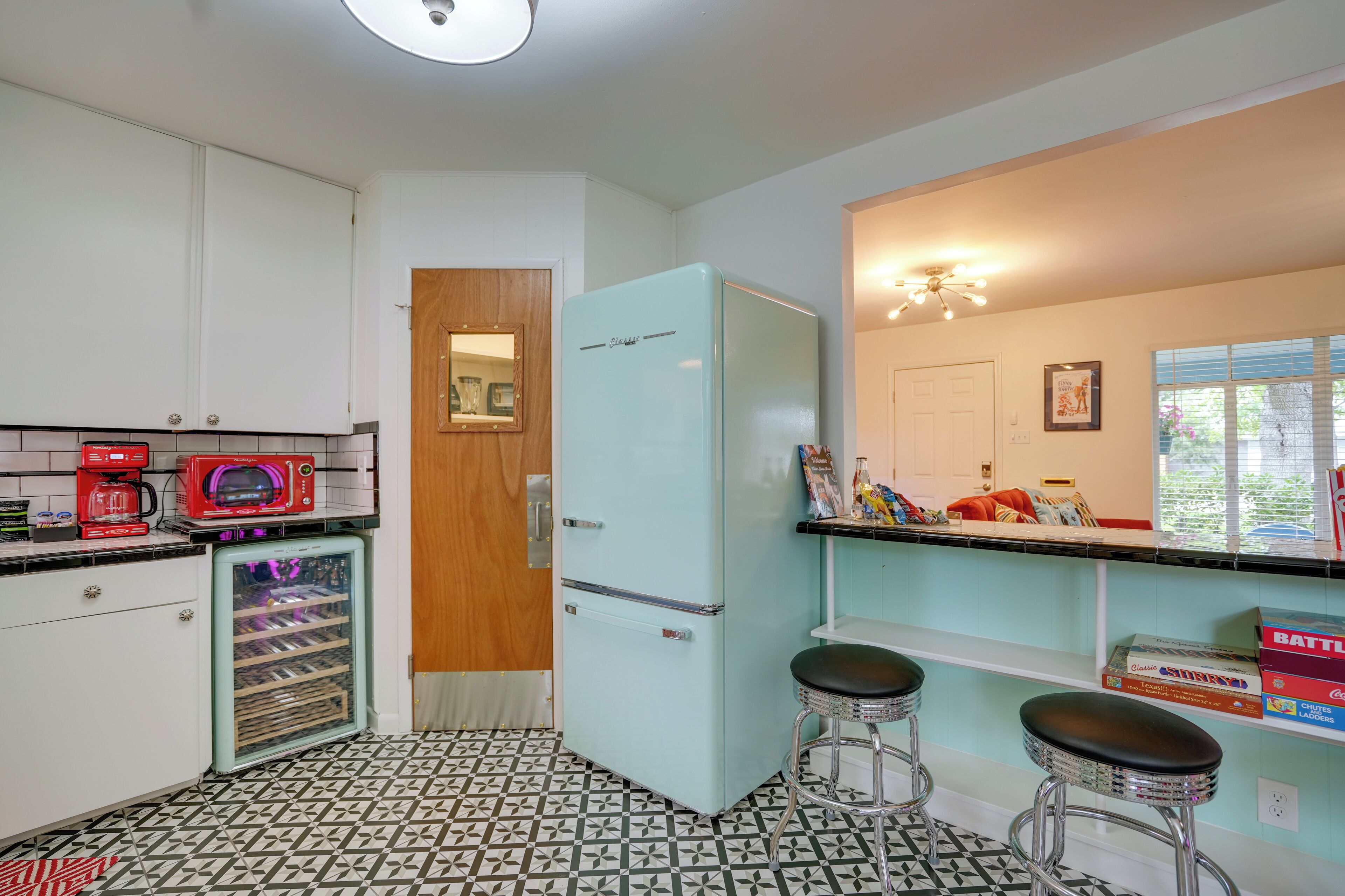 A kitchen with a light blue refrigerator, a red microwave, and a small refrigerator. There are two stools and a window with blinds.