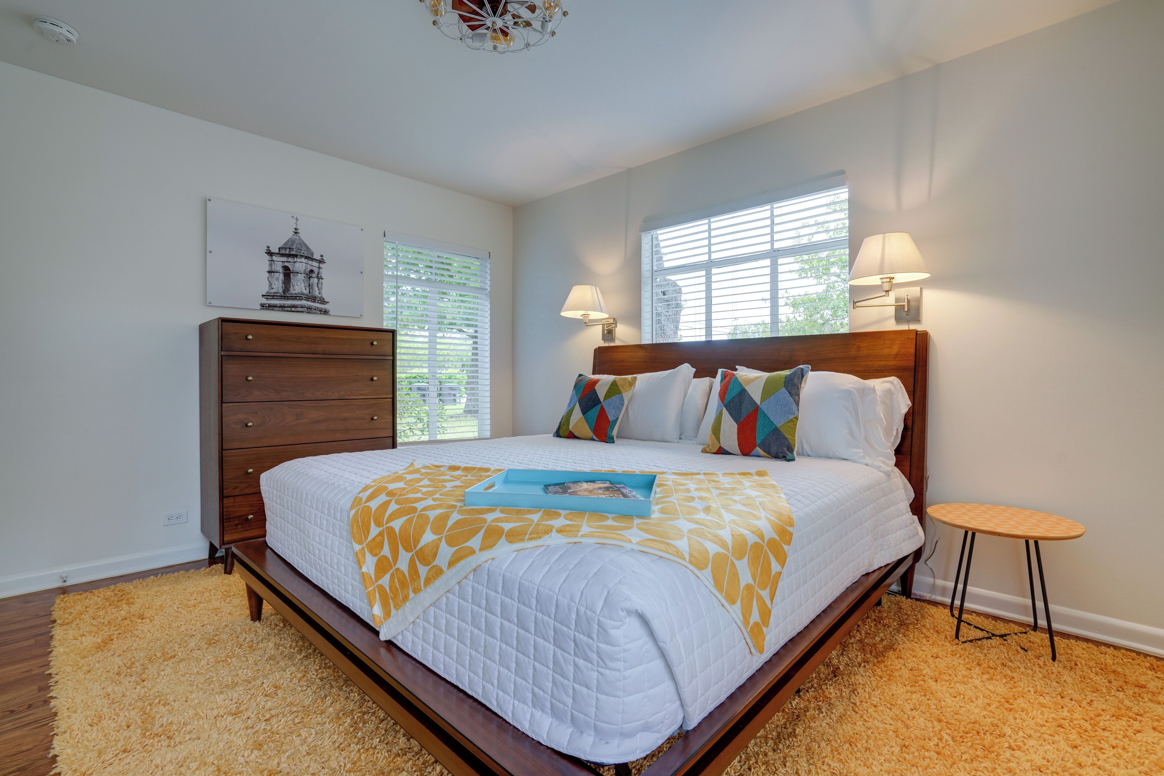 A bedroom with a wooden bed, a small round table, a dresser, and a window with blinds.