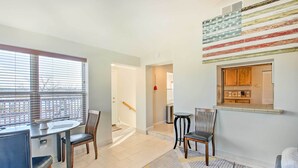 Una sala de estar moderna con comedor, una cocina visible a través de una ventana y una escalera que sube a otro nivel.