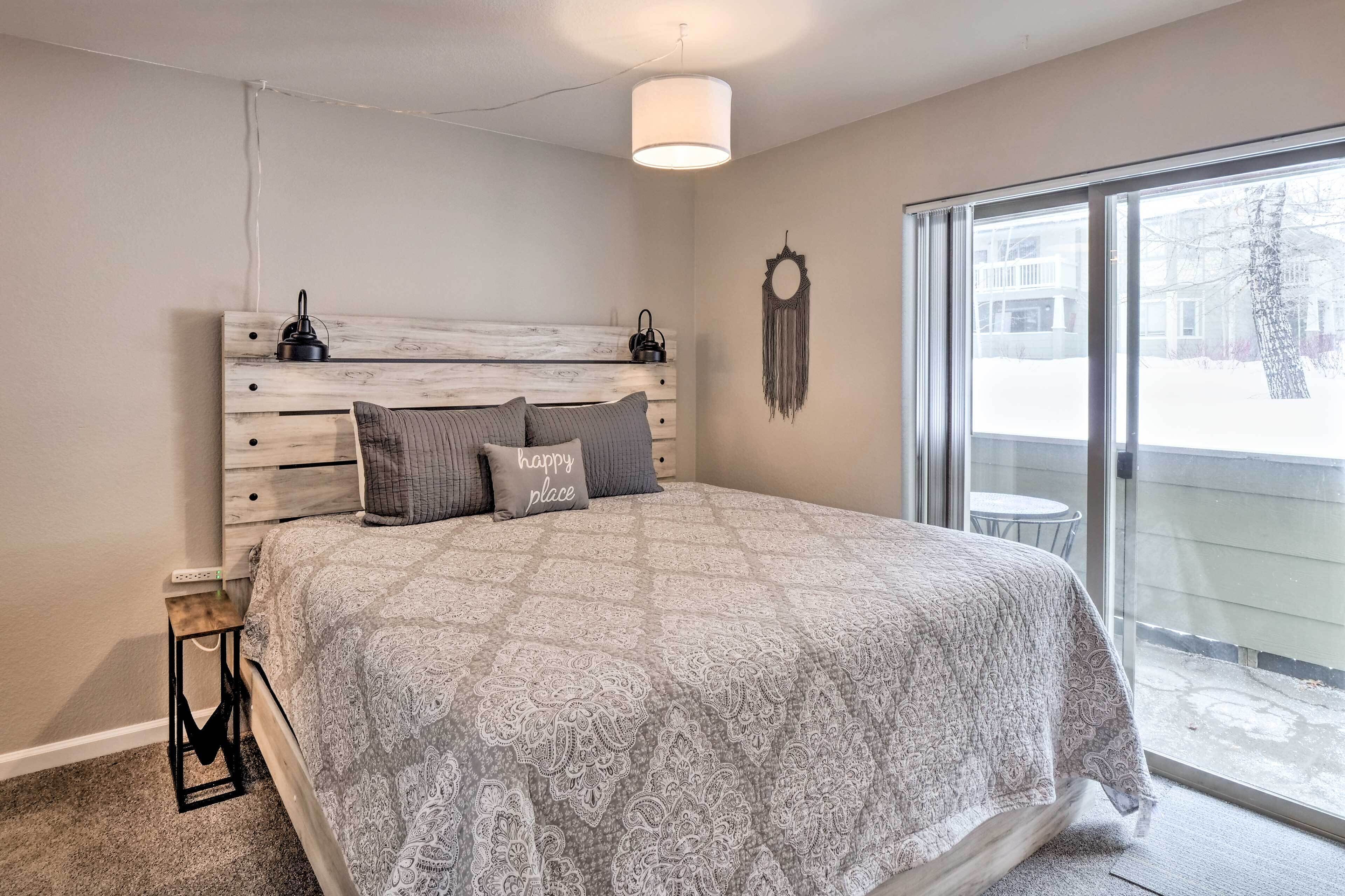 A bedroom with a large bed, a wooden headboard, a bedside table, a wall-mounted lamp, and a sliding glass door leading to a balcony.