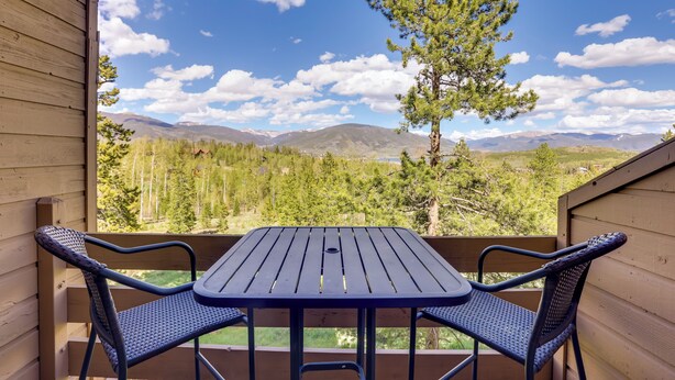 Eine Holzterrasse mit Tisch und zwei Stühlen, mit Blick auf eine Berglandschaft mit Bäumen und blauem Himmel.