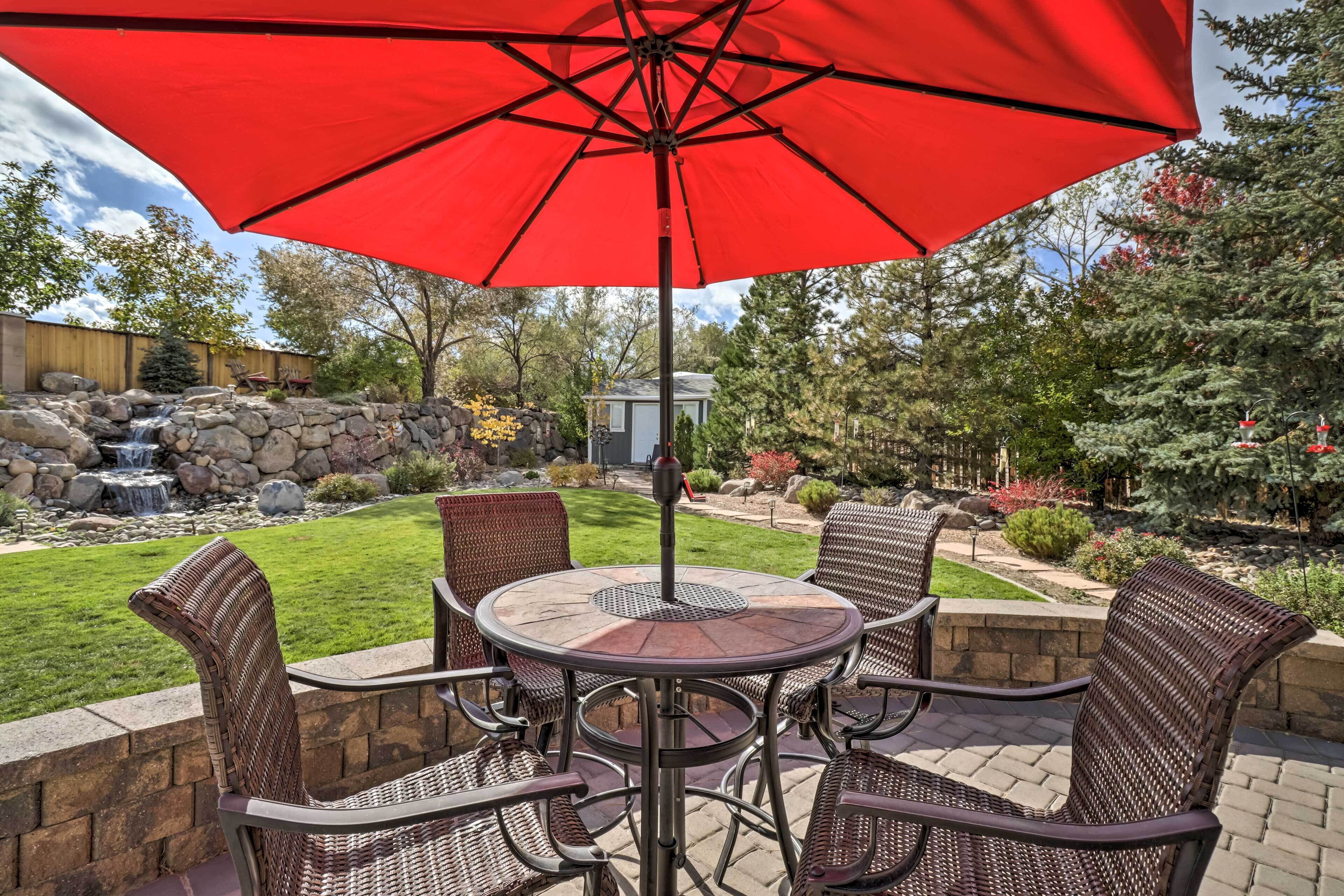 A patio with wicker chairs and a red umbrella, a stone wall, and a small waterfall in the background.