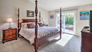A bedroom with a four-poster bed, a nightstand with a lamp, a dresser, and a large window with a view of greenery. - Large Yard + Hot Tub: Reno Retreat! (Reno)