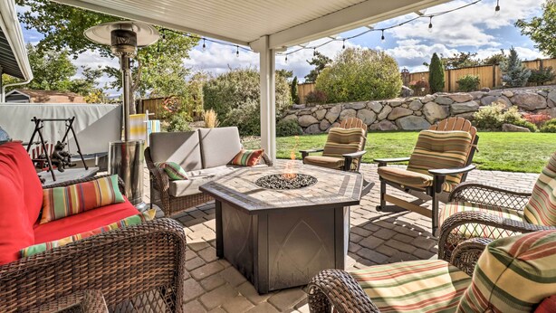 A covered patio area with wicker furniture, a stone fire pit, and a view of a backyard with a stone wall and greenery. - Large Yard + Hot Tub: Reno Retreat! (Reno)
