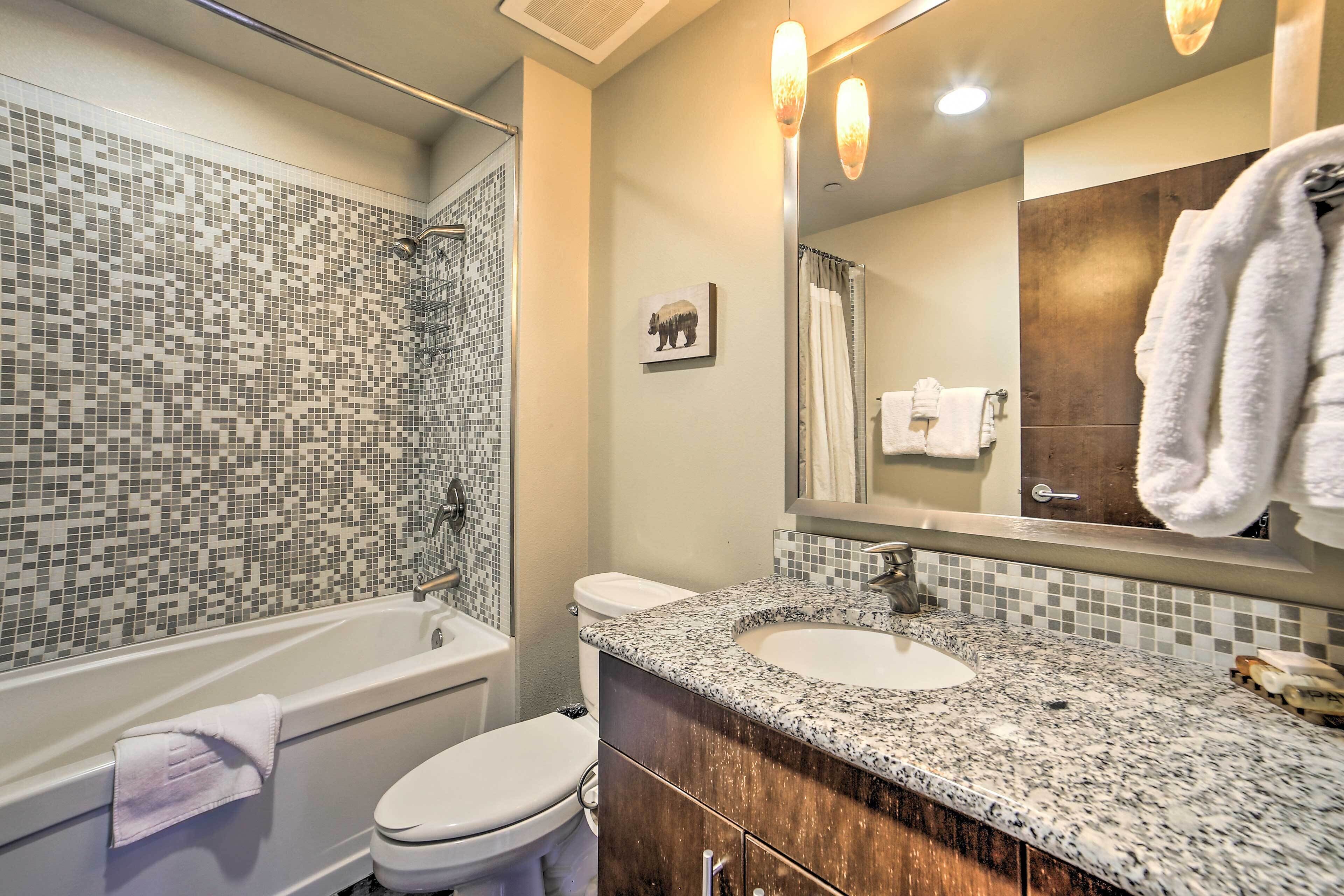 A bathroom with a mosaic tile shower, a white toilet, a sink with a granite countertop, and a mirror with a towel hanging on it.