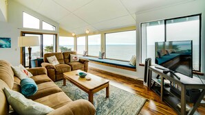 A spacious living room with a large window seat overlooking the sea, a television, and a wooden coffee table.