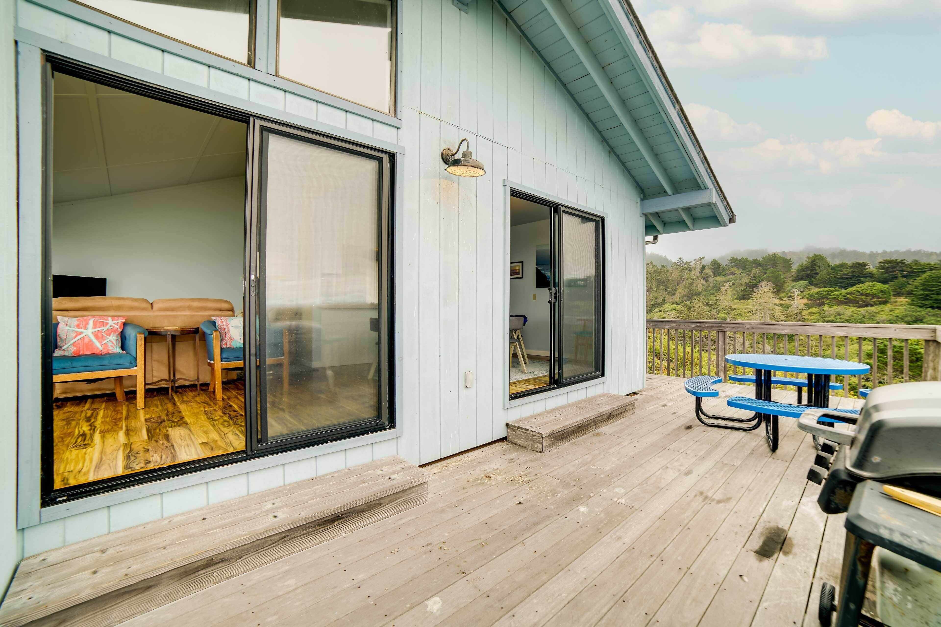 A wooden deck with a barbecue grill, a covered seating area with blue cushions, and a view of the surrounding landscape.