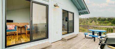 A wooden deck with a barbecue grill, a covered seating area with blue cushions, and a view of the surrounding landscape.
