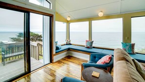 A room with large windows overlooking a body of water, a wooden deck outside, and a seating area with a sofa and armchairs.