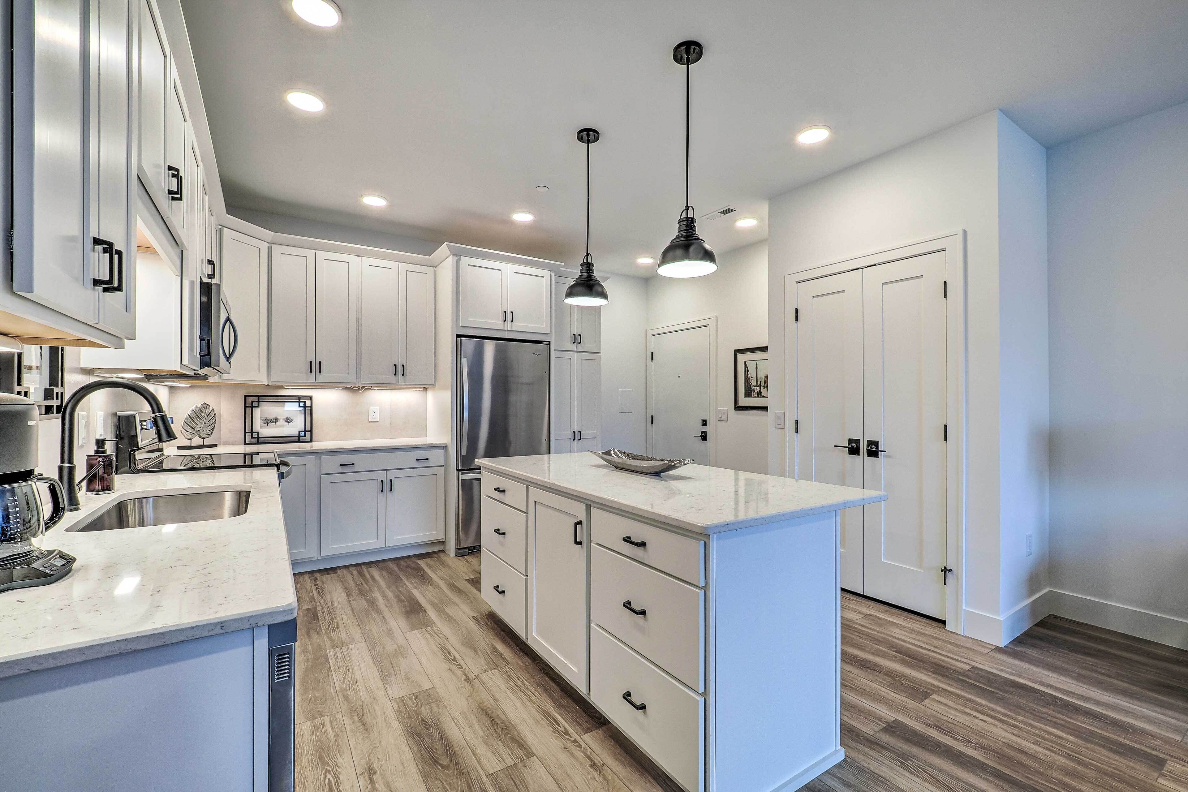 A modern kitchen with white cabinets, a stainless steel refrigerator, a large island with a bowl, and pendant lighting.