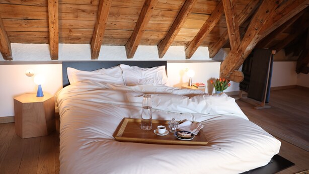 A bedroom with a wooden ceiling, a bed with white linens, a bedside table with a lamp, and a tray with a bottle, cup, and flowers. - Sessius 1622 (Cessy)