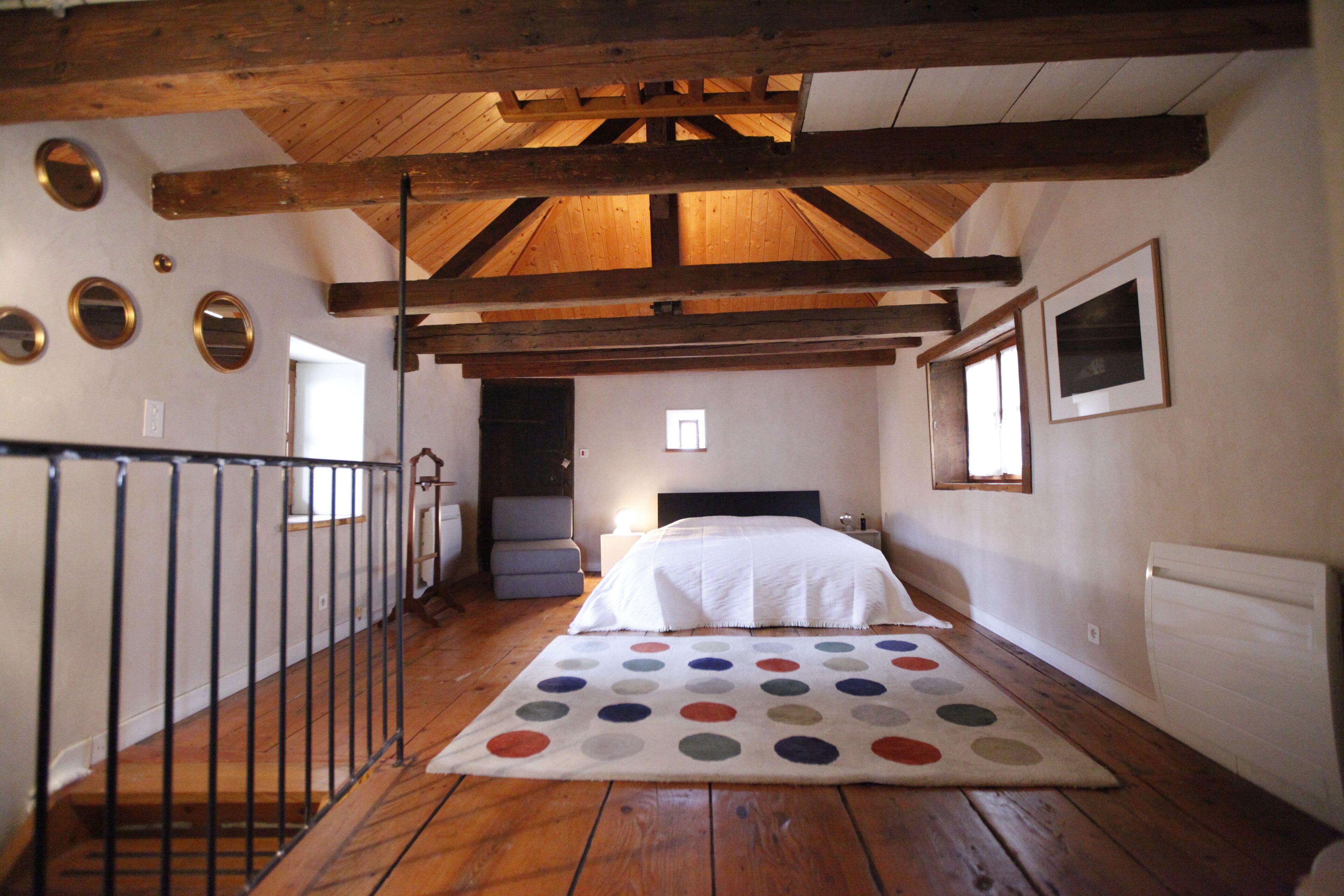 A bedroom with a wooden ceiling, a bed with a white bedspread, a polka dot rug, and a small window.