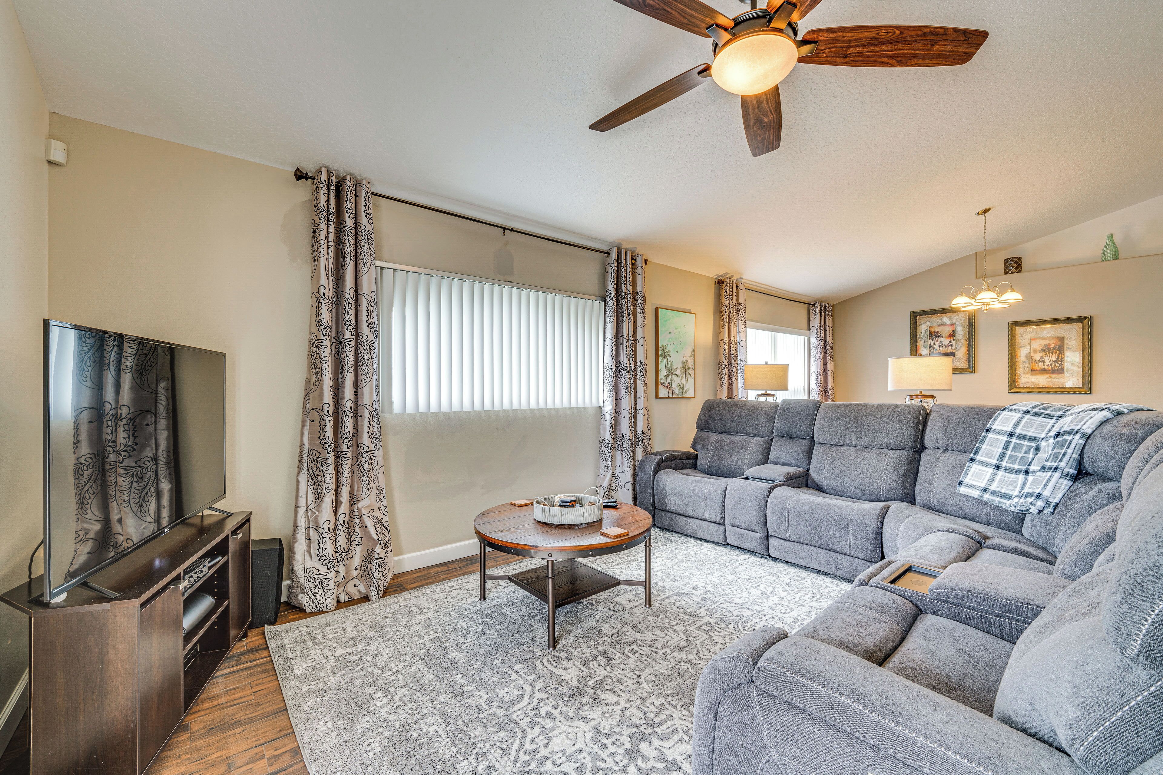 A living room with a flat-screen TV, a ceiling fan, a gray sectional sofa, and a patterned rug.