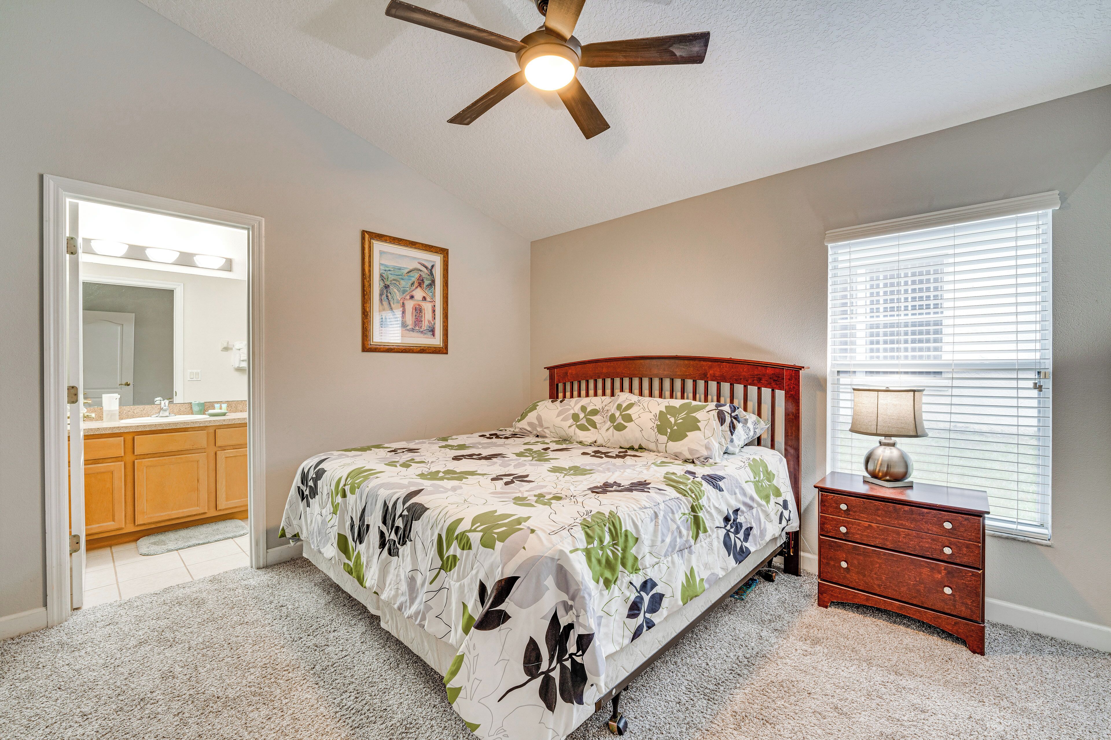 A bedroom with a bed, a nightstand, a ceiling fan, a window with blinds, and a door leading to a bathroom.