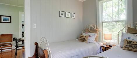 Une chambre avec deux lits, un banc en bois, une chaise et une fenêtre avec des stores.