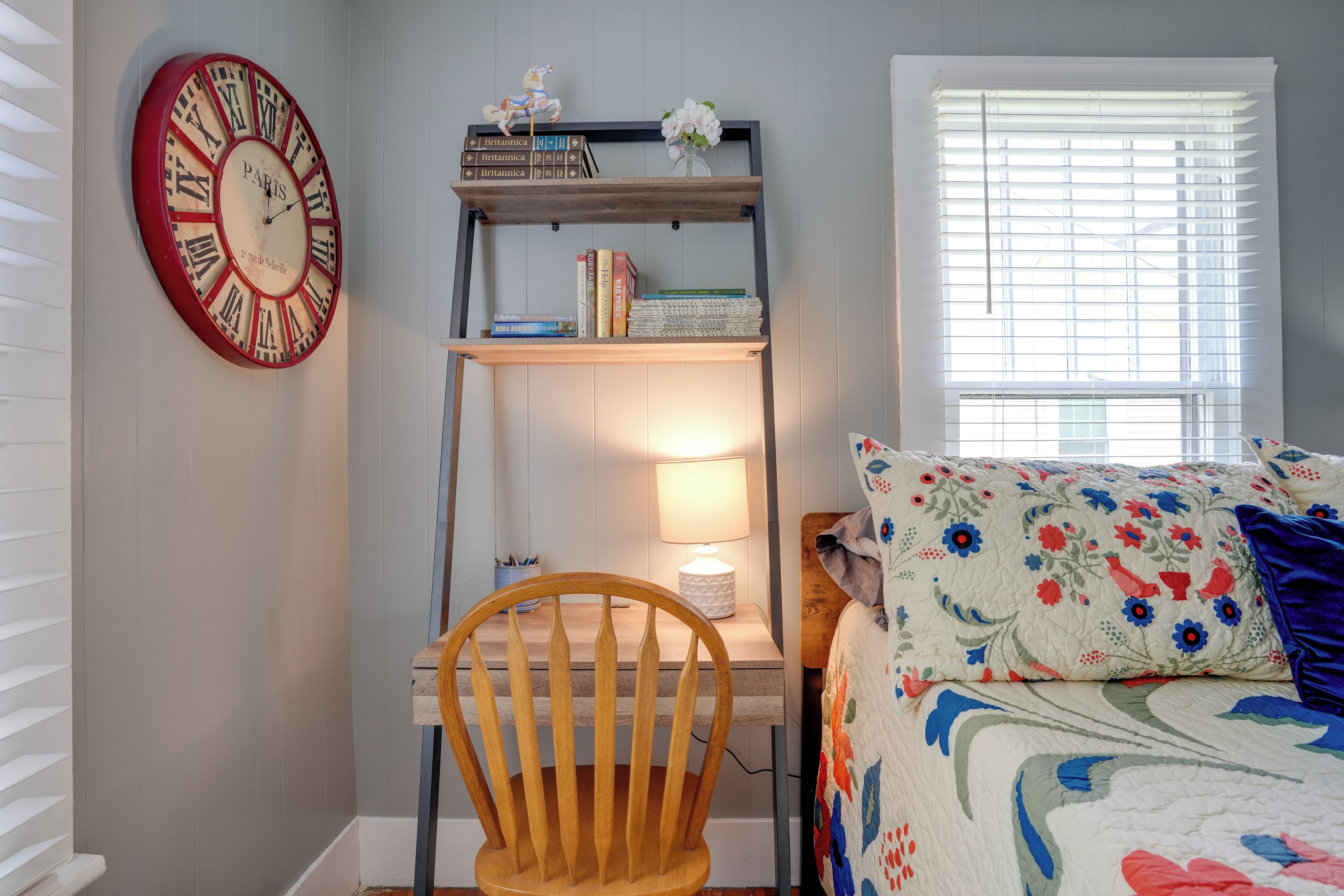 Une chambre avec une chaise en bois, une bibliothèque remplie de livres, une horloge décorative et une fenêtre avec des stores.