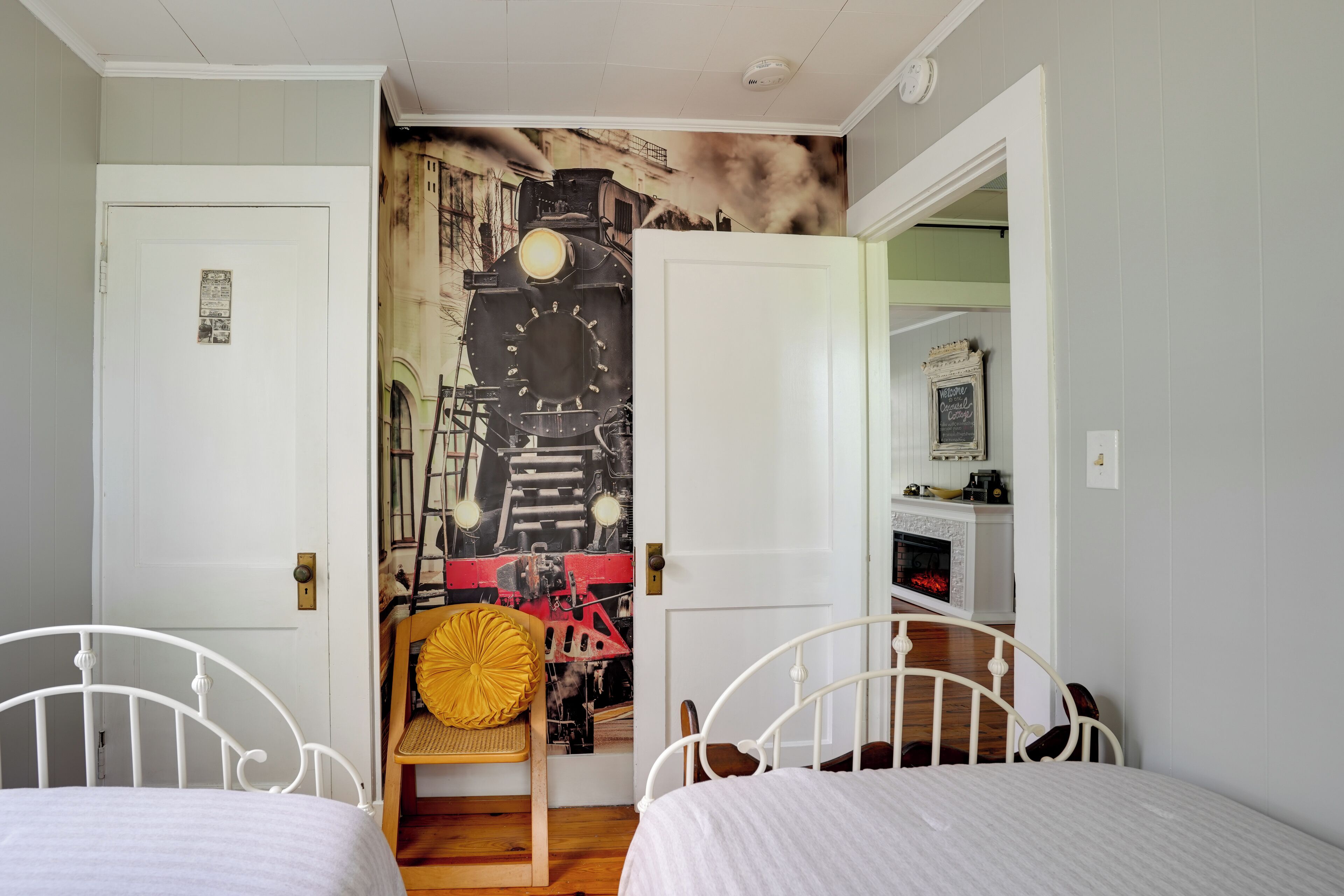 Un dormitorio con dos camas individuales, una silla de madera, un mural en la pared y una pequeña zona de cocina.