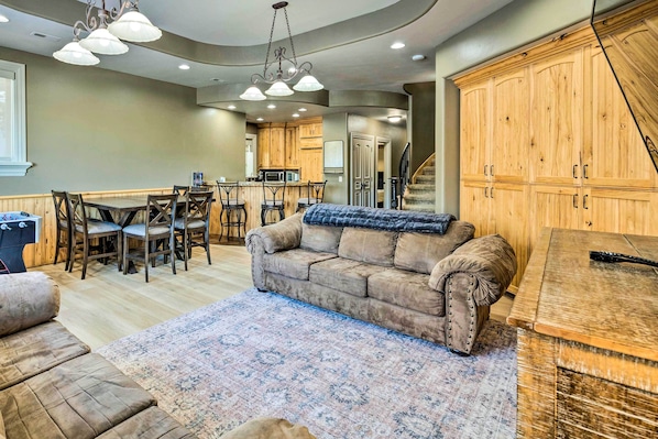 A spacious living area with a large couch, a coffee table, and a patterned rug. There is a kitchen in the background with wooden cabinets and a dining area with a table and chairs. - Family-friendly Utah Vacation Rental Near Skiing! (Sandy)