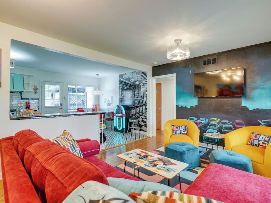 Un salón con un sofá rojo, sillones amarillos y una mesa de centro. Se ve una cocina al fondo con un jukebox.