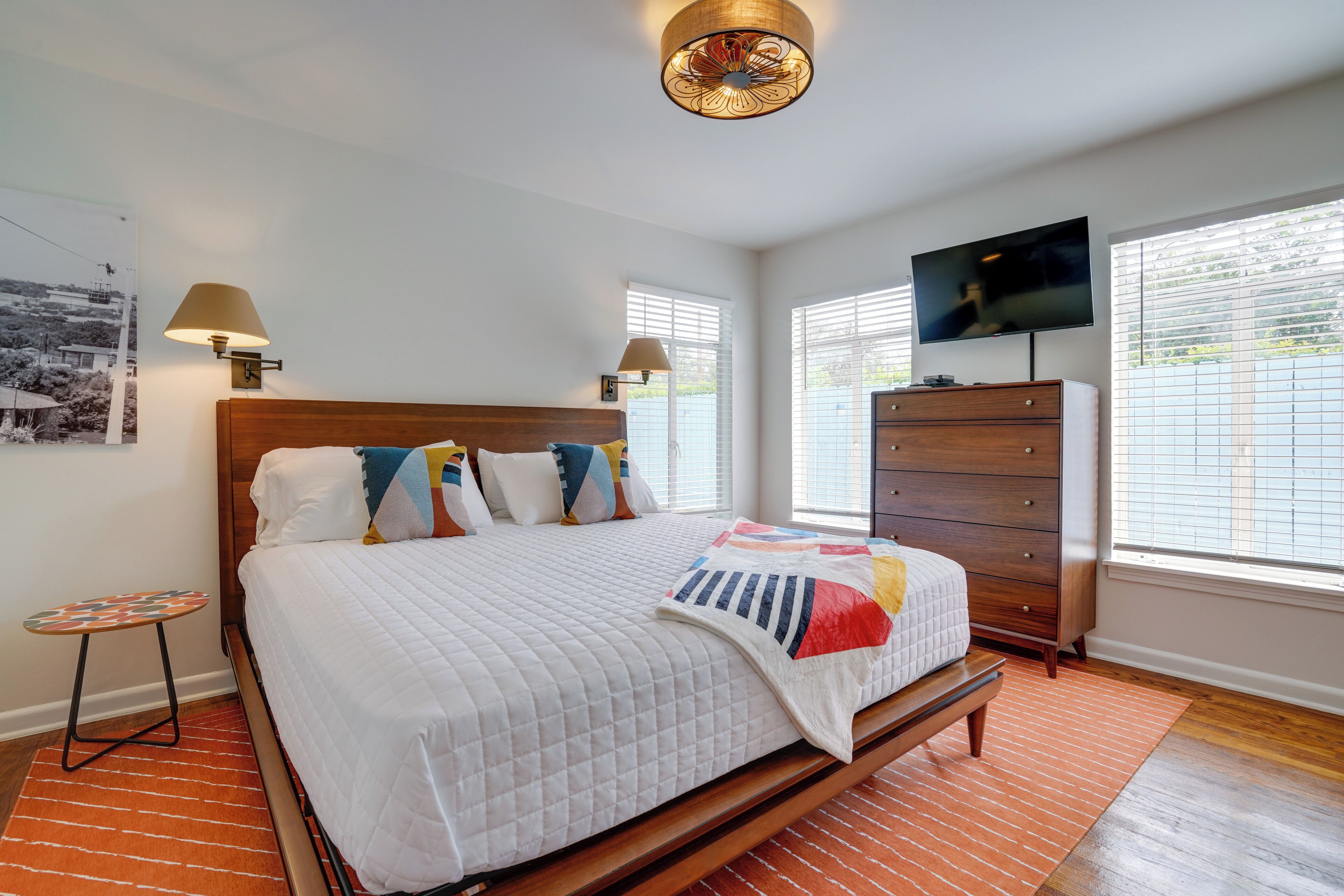 A bedroom with a wooden bed, two bedside lamps, a dresser, a television, and a window with blinds.
