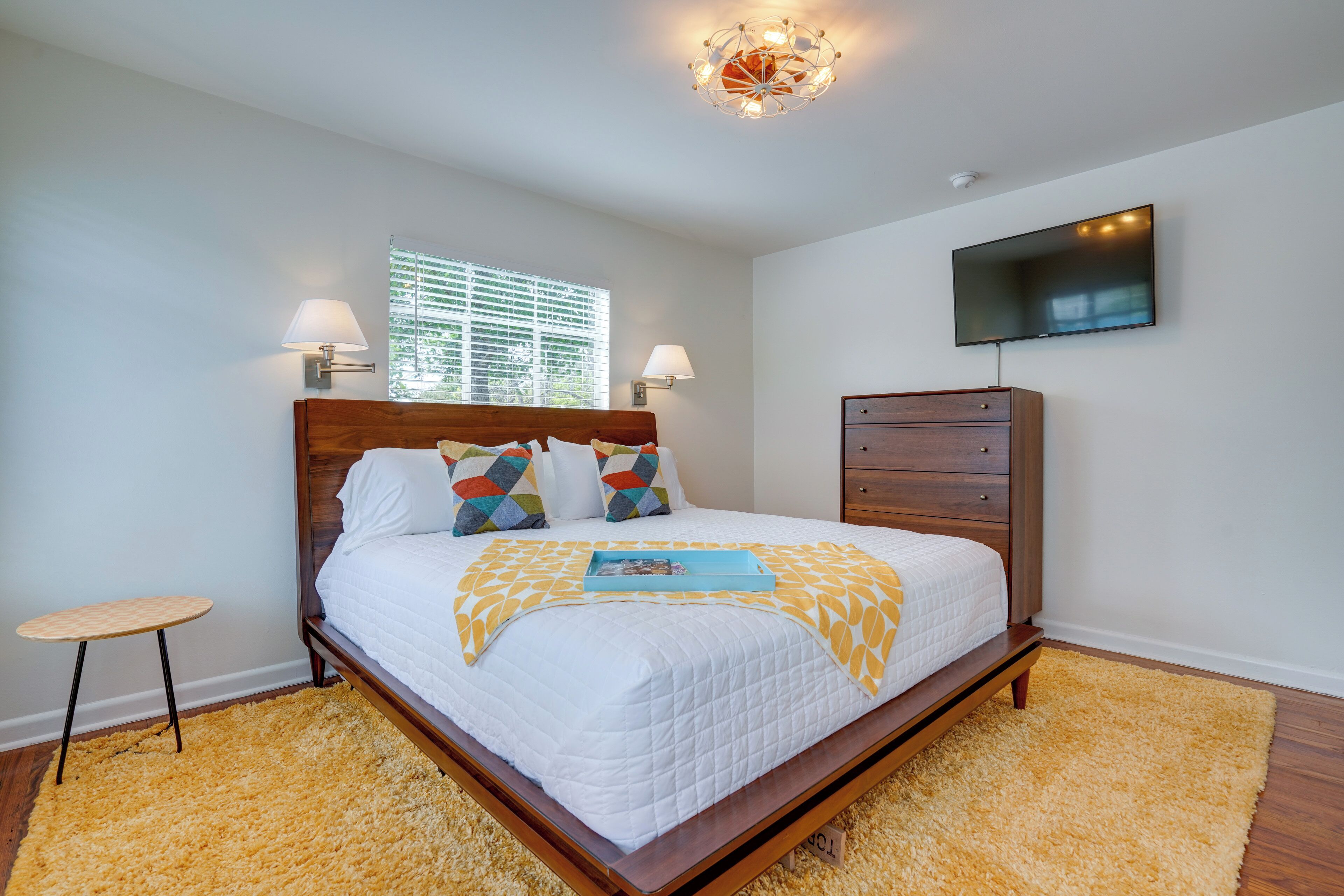 A bedroom with a wooden bed, a small bedside table, a TV on the wall, and a window with blinds.