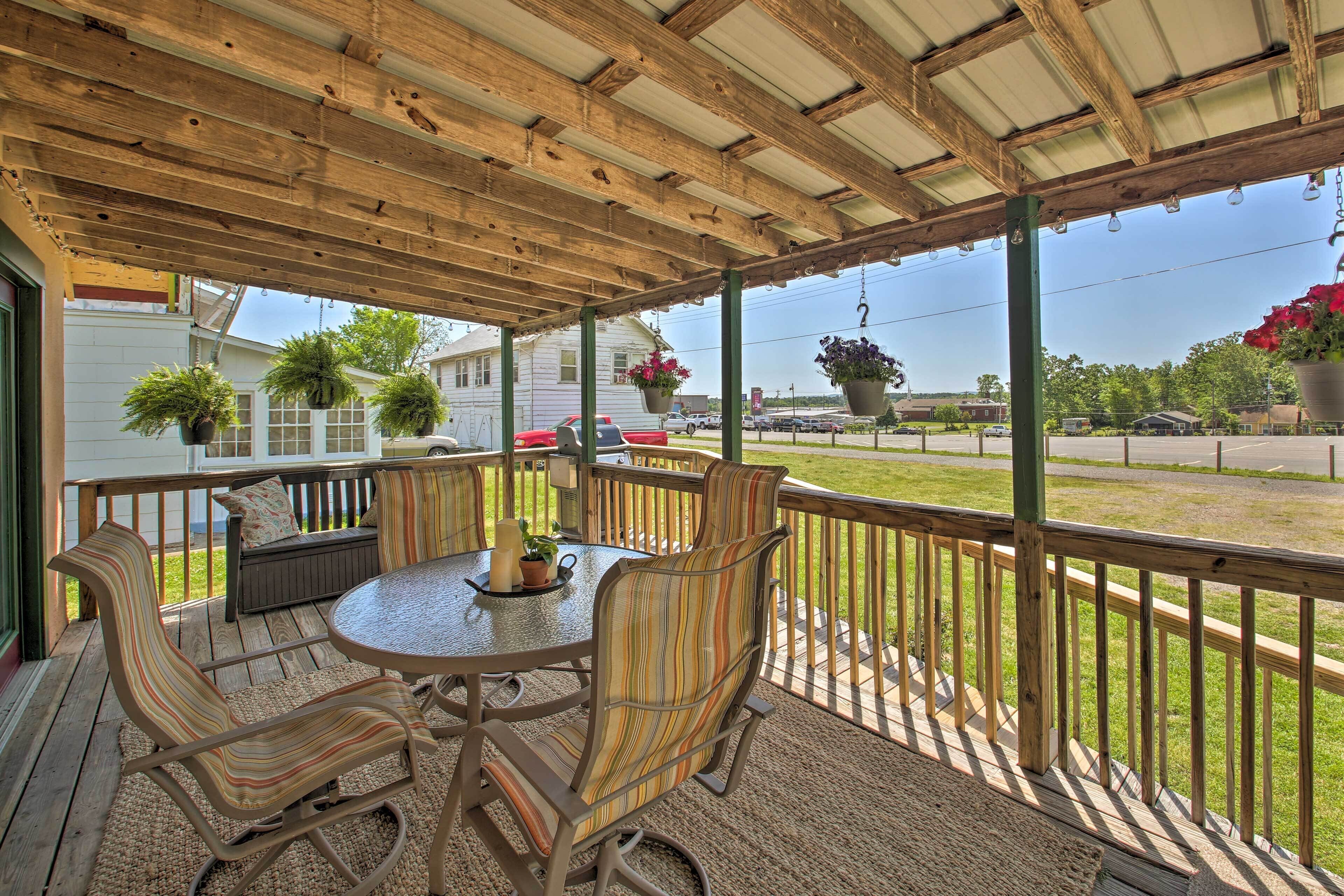 A covered patio with a round table and striped chairs, surrounded by hanging planters and a view of the street.