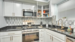 Een moderne keuken met witte kasten, een ingebouwde magnetron, fornuis en oven. Er is een granieten aanrecht, een spoelbak met kraan en een raam met zonwering.