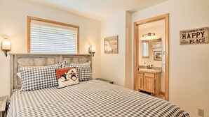 A bedroom with a checkered bedspread, a wooden headboard, a window with blinds, a wall-mounted light, a framed picture, and a door leading to a bathroom with a wooden cabinet and a mirror.