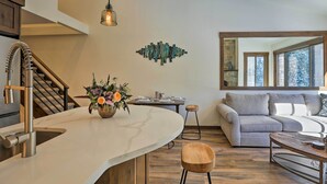 Une cuisine moderne dotée d’un îlot central aux lignes courbes, d’un escalier en bois, d’un coin repas avec une table et des chaises, et d’un salon avec un canapé et une table basse.