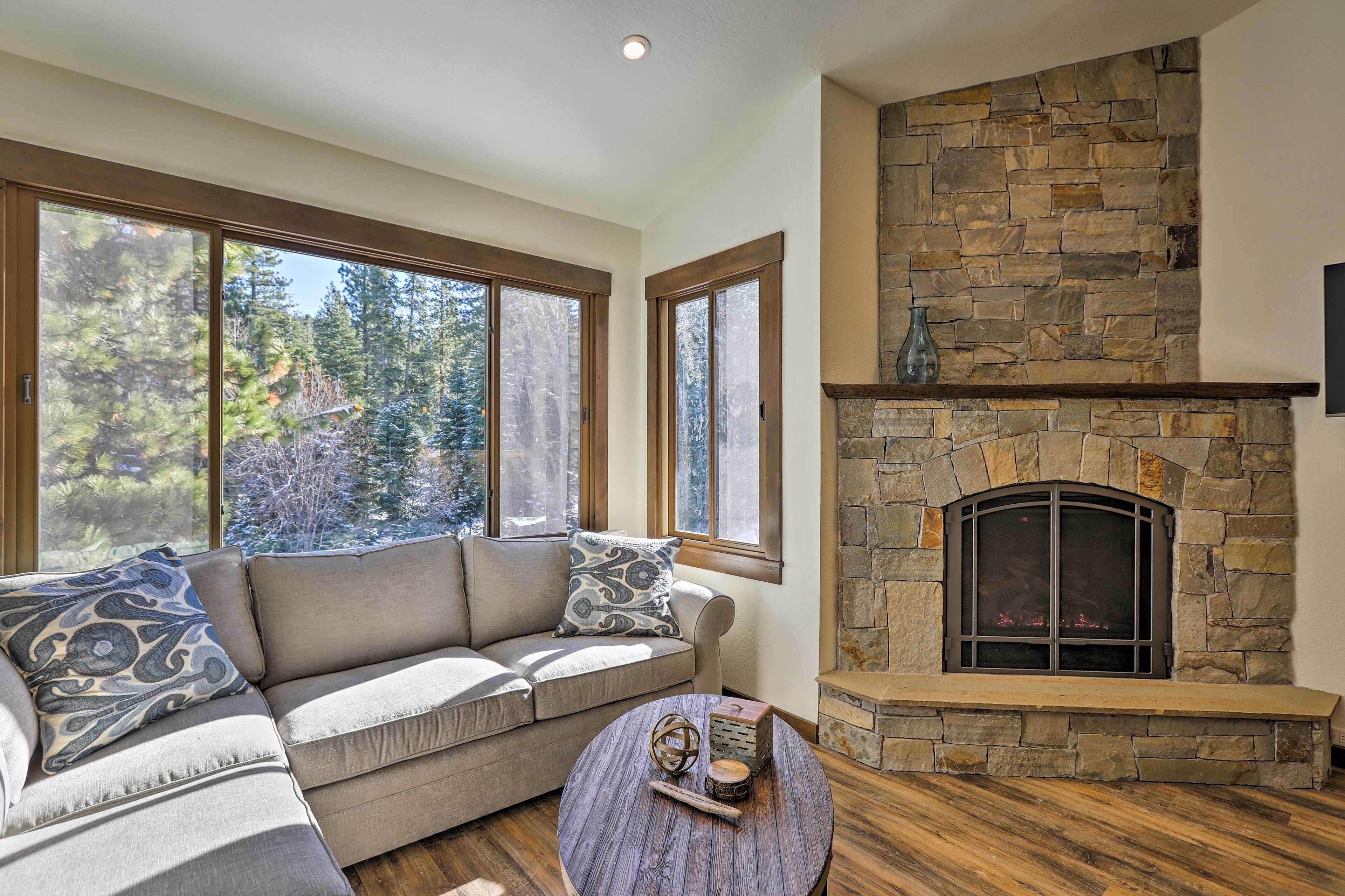 A cozy living room with a stone fireplace, a beige sofa with patterned cushions, and large windows offering a view of trees.