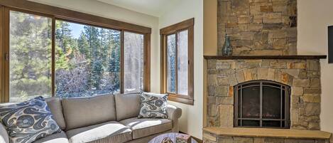 A cozy living room with a stone fireplace, a beige sofa with patterned cushions, and large windows offering a view of trees.