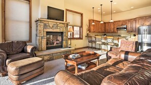 A living room with leather furniture, a stone fireplace, wooden cabinets, and a kitchen area with stainless steel appliances. - Crested Butte Townhome w/ Views - Steps to Lifts! (Crested Butte)