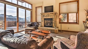 A spacious living room with a stone fireplace, leather sofas, and a large window offering a view of the outdoors. - Crested Butte Townhome w/ Views - Steps to Lifts! (Crested Butte)