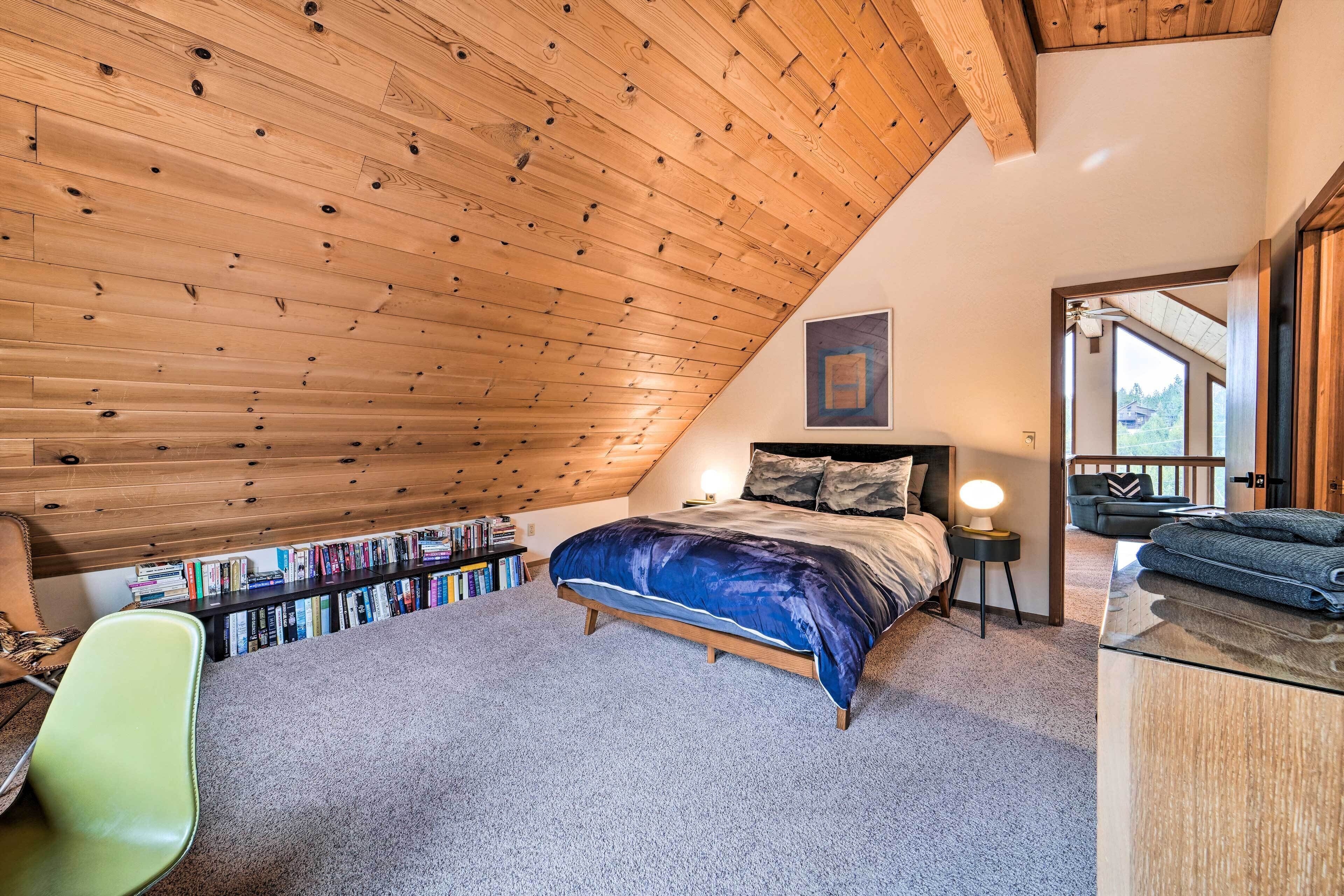 A bedroom with a sloped ceiling, a bed with a blue comforter, a bedside table with a lamp, a bookshelf, and a chair.