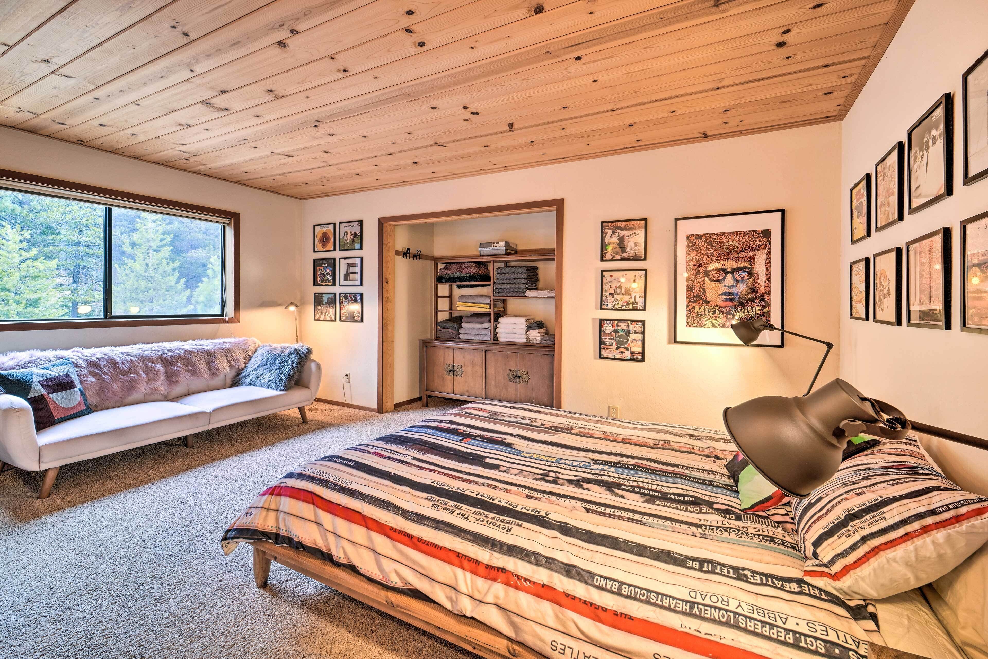 A cozy bedroom with a wooden ceiling, a large window, a bed with a striped comforter, a sofa with a fur throw, and a bookshelf.