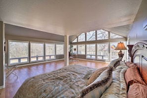 A spacious bedroom with large windows, a bed with patterned bedding, and a nightstand with a lamp. - Grand Tulsa Getaway With Private Pond, Dock & Deck (Tulsa)