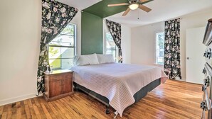 Un dormitorio con cama, piso de madera, paredes verdes, cortinas con estampado floral y un ventilador de techo.