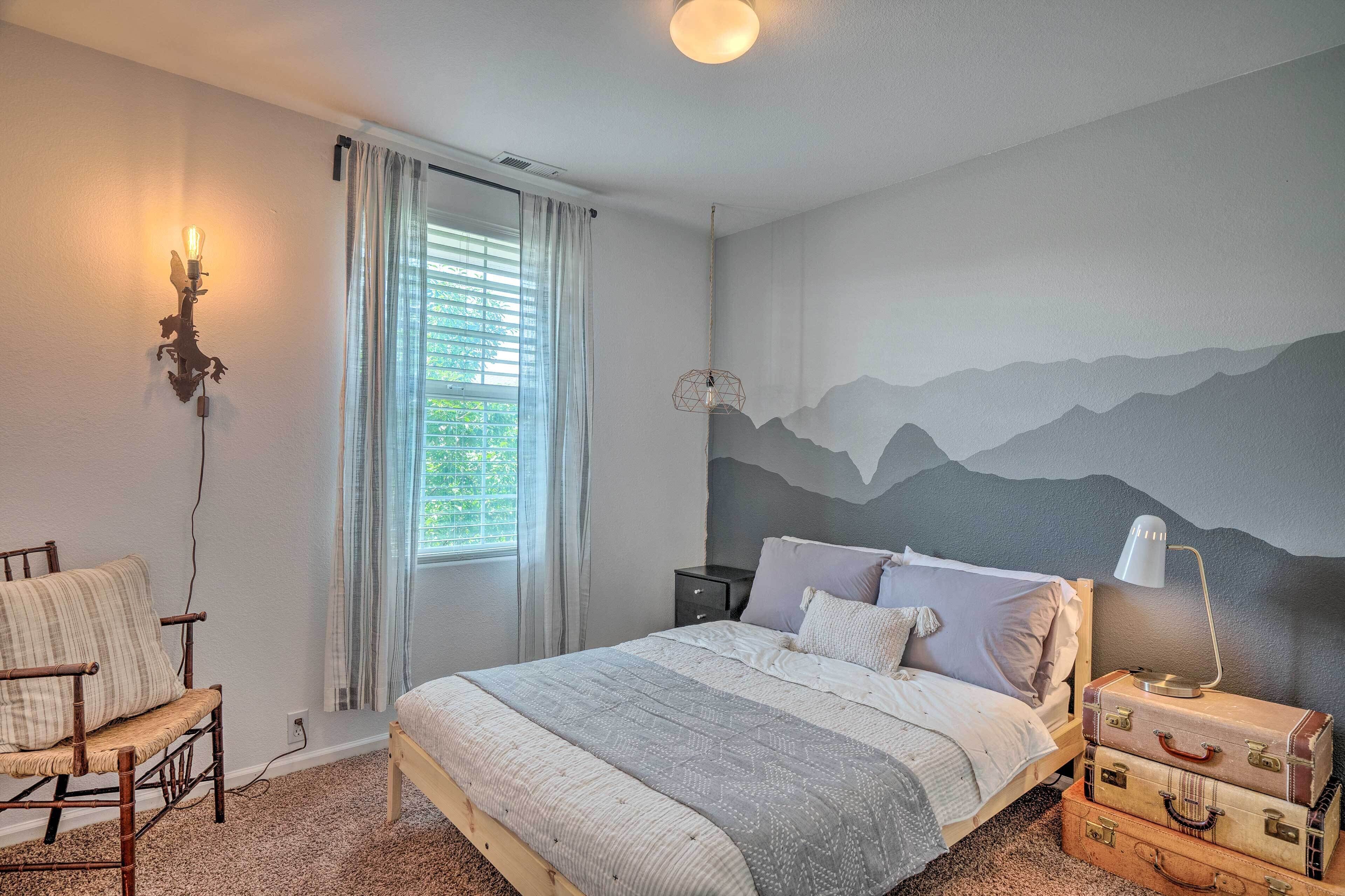 Un dormitorio con cama, una silla, una mesita de noche, una lámpara y un mural en la pared.